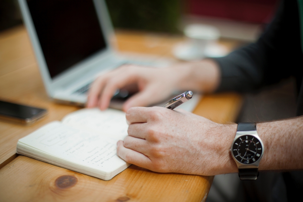 man writing in a notebook on a table with a laptop in the background