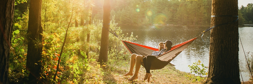couple in hammock out in the woods next to trees with a lake behind the,