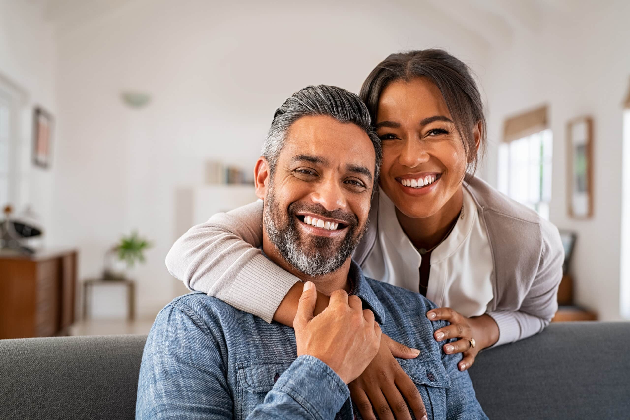 Smiling african american woman hugging mid adult man sitting on couch from behind at home.
