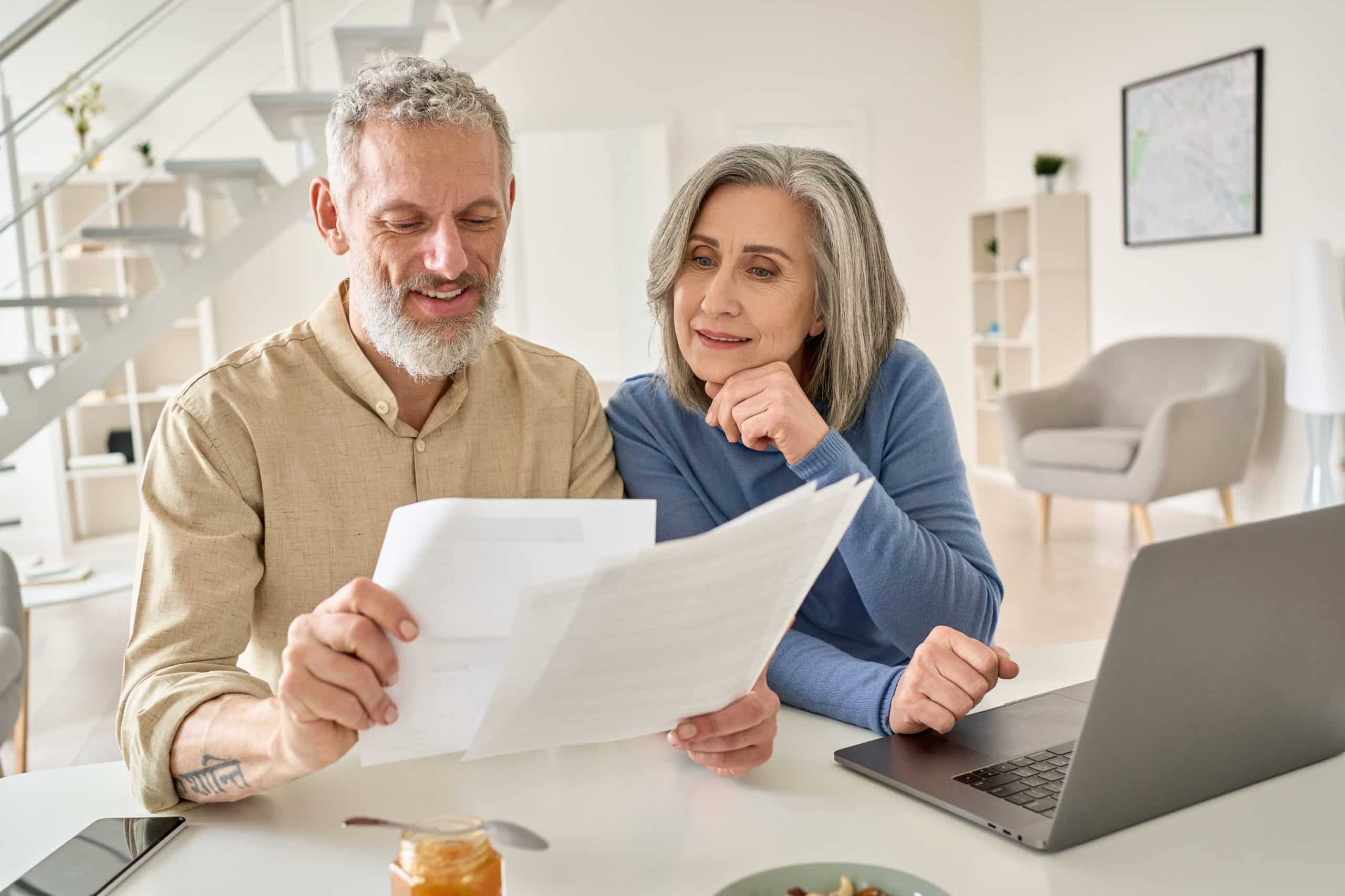 Middle aged couple reading paper bills calculating pension using laptop at home