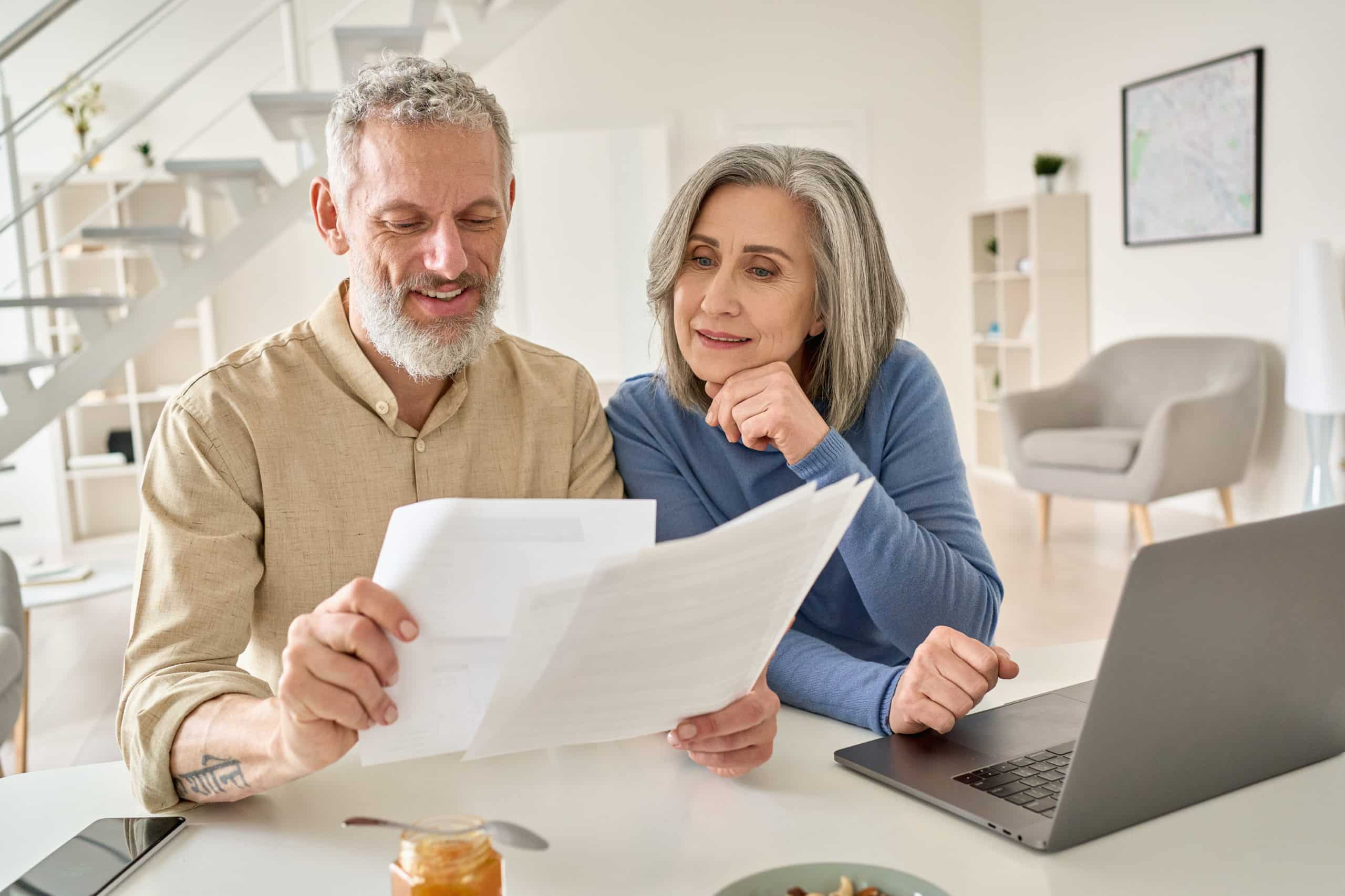 Middle aged couple reading paper bills calculating pension using laptop at home